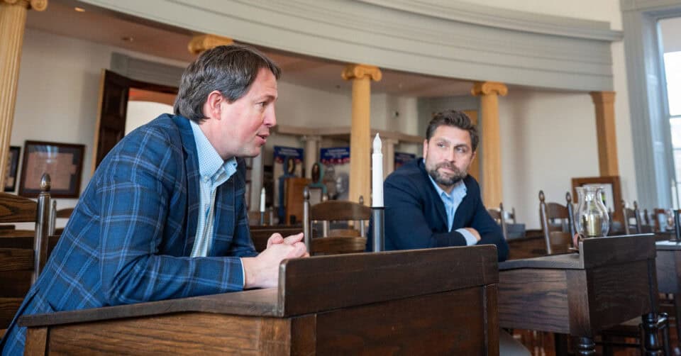 Two men seated at wooden tables.